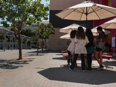 A small group of students stand underneath a table umbrella that provides some shade in a schoolyard during a hot day.