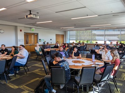 People sit in round-shaped tables with voting information materials in a conference room at a university.