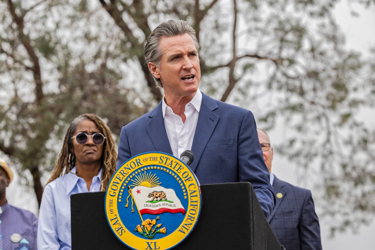 Gov. Gavin Newsom, wearing a blue suit and white shirt, stands behind a black podium with the California state seal on it during a press conference.