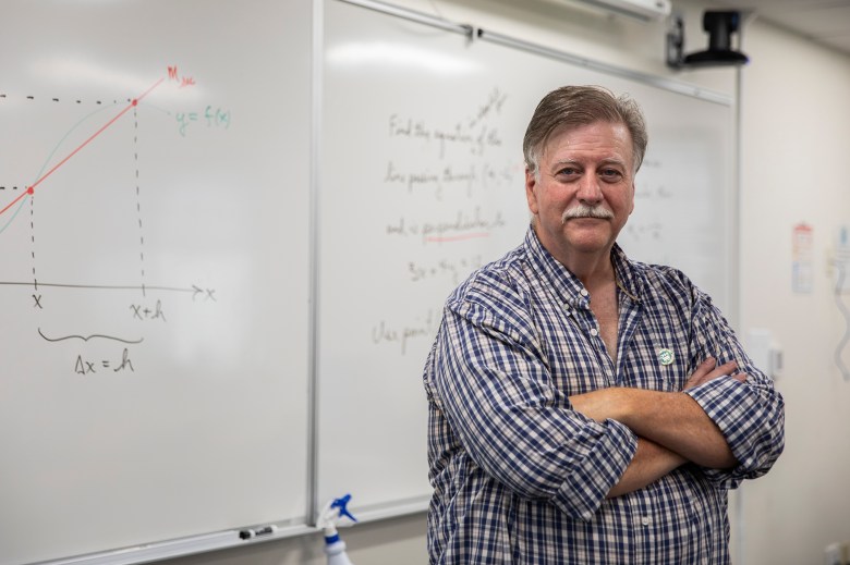 A man standing with his arms crossed and wearing a faculty pin on his buttoned-up shirt in front of a whiteboard with equations written on it.