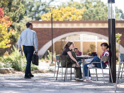 A man with a brief case walking pass two young women sitting at a table talking at a college campus.