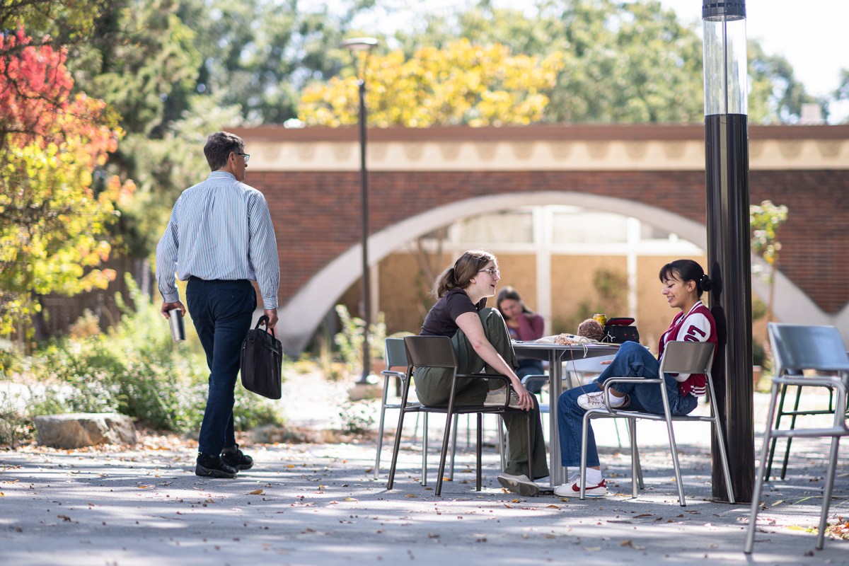 A man with a brief case walking pass two young women sitting at a table talking at a college campus.