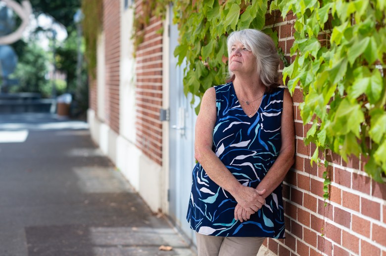 A woman is leaning against a brick wall with vegetation growing along the wall.