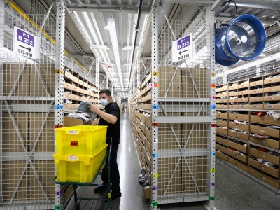 A person wearing a face mask works in a large warehouse aisle lined with shelves filled with boxes. They are sorting items from a cart holding two bright yellow bins labeled "HEAVY." Overhead, fluorescent lights illuminate the space, and a large blue industrial fan is mounted on the right side. Signs on the shelves indicate section numbers and rows.