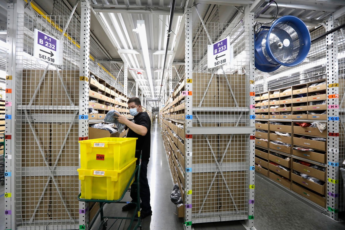 A person wearing a face mask works in a large warehouse aisle lined with shelves filled with boxes. They are sorting items from a cart holding two bright yellow bins labeled "HEAVY." Overhead, fluorescent lights illuminate the space, and a large blue industrial fan is mounted on the right side. Signs on the shelves indicate section numbers and rows.