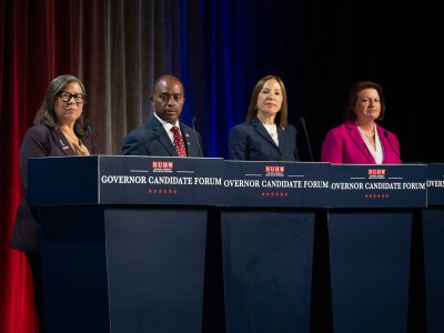 Four people stand on a stage behind individual podiums, each looking towards their right in front of an audience at a governor candidate forum. Blue, red and gray lights illuminate the stage curtains the background.