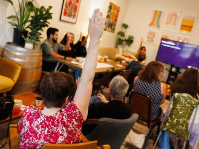 A woman wearing a red pattern shirt raises her among a room with attendees at a voter information event.