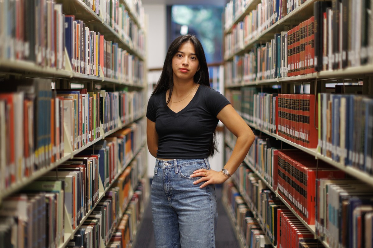 A person stands in the middle of a library aisle, surrounded by shelves filled with books. They are wearing a black V-neck shirt and blue jeans, with one hand on their hip and the other relaxed by their side. The background shows books neatly organized on both sides, leading to a window at the far end.