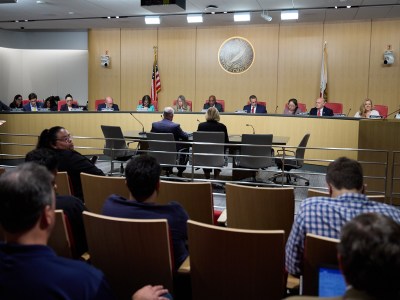 People sit at a panel table in front of microphones, speaking to an audience during a hearing.