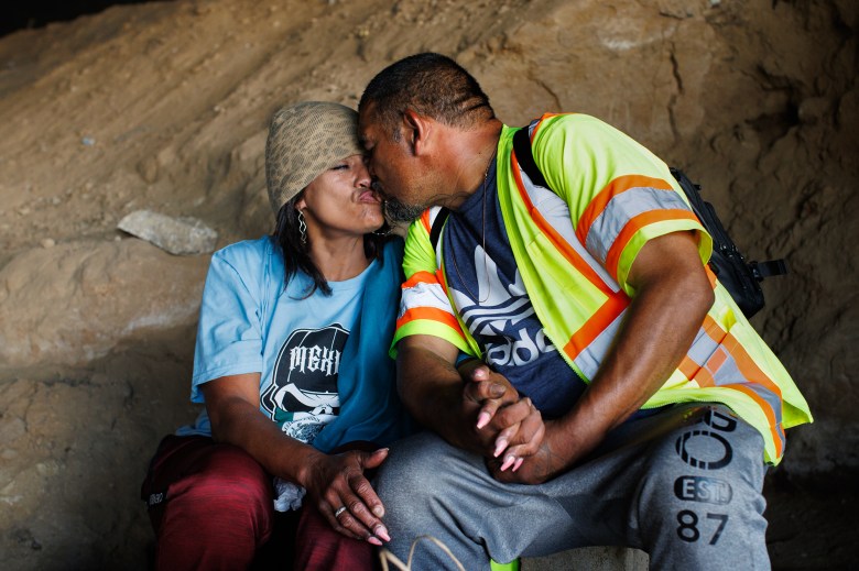 Two people kiss on the lips as they hold hands and sit next to each other in a cove underneath a freeway. The person on the left wears a brown beanie, blue graphic t-shirt and red pants. The person on the right wears a safety vest, graphic t-shirt and grey pants.