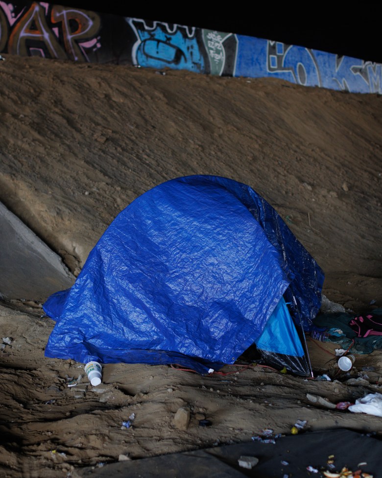 A tent with a blue tarp on top sits in a dirt area underneath a freeway bridge with trash around it.