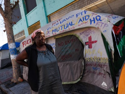 A person wearing a pink leopard-patterned head wrap stands in front of a sidewalk with tents of homeless residents as her arms rest on her back.