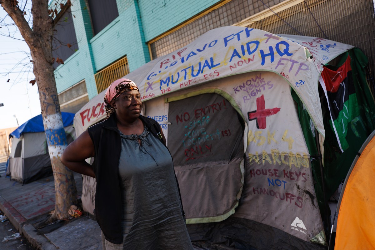 A person wearing a pink leopard-patterned head wrap stands in front of a sidewalk with tents of homeless residents as her arms rest on her back.