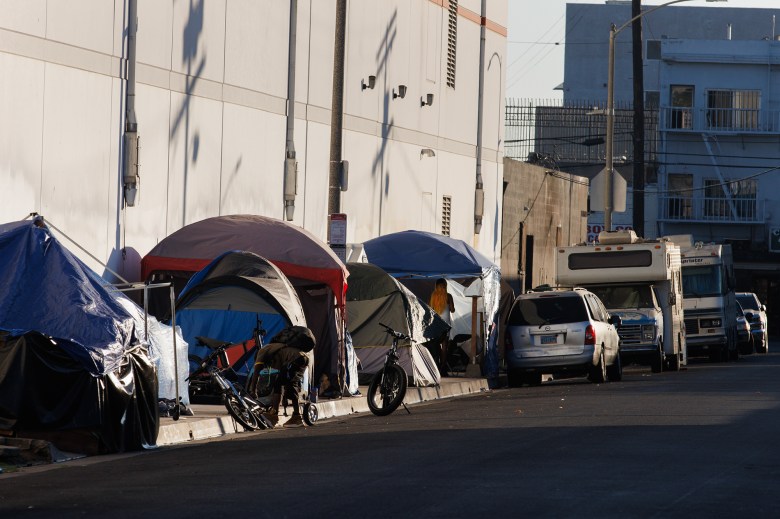 A side view of an afternoon sun shining on a street lined with tents for those experiencing homelessness with some cars and bikes nearby.