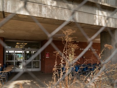 The entrance to "Davies Hall," a building partially obscured by a metal chain-link fence. The foreground features dry plants and vegetation, which are visible through the fence. Outside the entrance, several blue chairs and desks are arranged. The structure of the building above the entrance is made of concrete, and the name "Davies Hall" is visible on the wall to the right of the doors. The overall atmosphere feels abandoned or under construction.