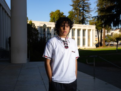 A person, wearing a traditional Mexican guayabera shirt and with their hands inside the pockets of their dark slack pants, stands in front of a building at a high school.