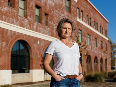 A person stands confidently in front of a red brick building, hands resting in their pockets. They are wearing a white T-shirt and jeans, with a determined expression as they look into the distance, sunlight highlighting their face. The building's arched windows and textured facade form an urban backdrop