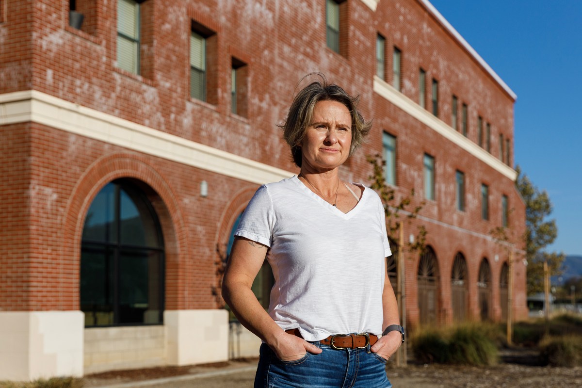 A person stands confidently in front of a red brick building, hands resting in their pockets. They are wearing a white T-shirt and jeans, with a determined expression as they look into the distance, sunlight highlighting their face. The building's arched windows and textured facade form an urban backdrop