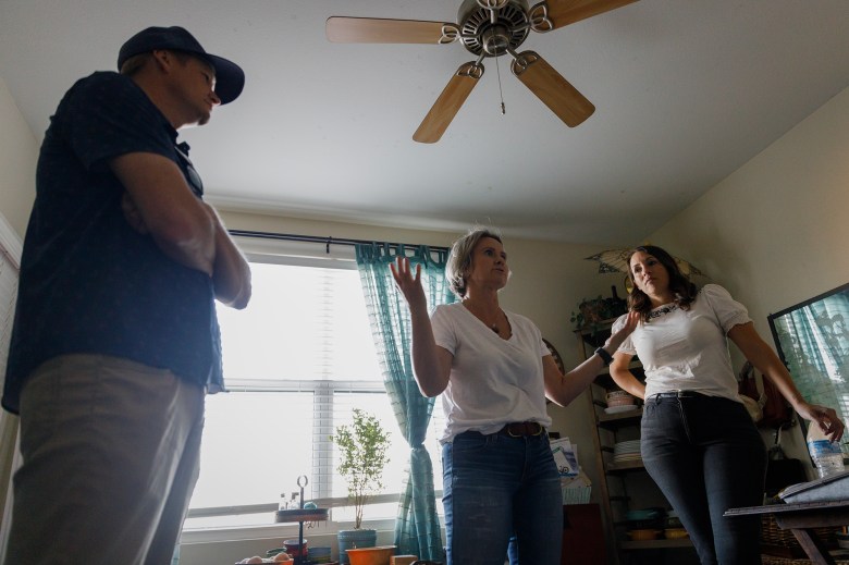 Three people are gathered in a room with one individual gesturing expressively. They stand near a window with teal curtains, partially illuminated by natural light. The other two individuals listen attentively, one with arms crossed and the other with a thoughtful expression. The ceiling fan above and shelves filled with personal items in the background add a homey atmosphere to the space.