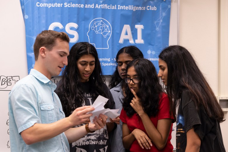 A teacher looking over pieces of papers with a group of students in front of a banner for the AI club in a classroom.