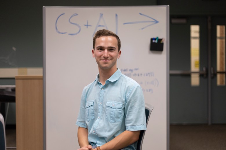 A man in a button up shirt smiles and sits in front of a white board with the AI club name on it.