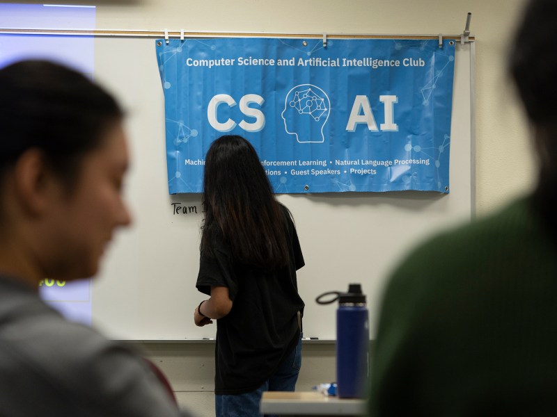 A student writing on a white board under a banner for the AI club in a classroom.