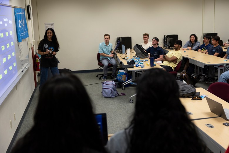 A group of students sitting in a row of tables inside of a classroom while another student stands at the whiteboard.