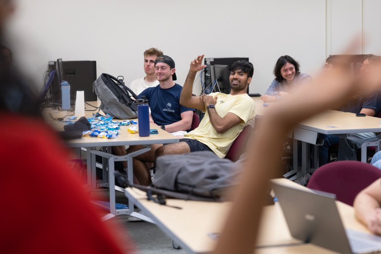 A classroom setting with several people seated around tables, some interacting and smiling. One individual in a yellow shirt raises a hand, appearing engaged in the discussion, while others look on with interest. The tables are scattered with items like water bottles, backpacks, and snacks. The foreground is partially blurred, with an arm raised, suggesting active participation.