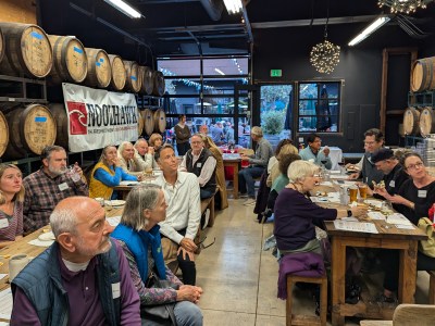 Attendees sit on wooden benches with drinks, food and flyers laid out on wooden tables in front of them as they listen to speakers at a brewing location with barrels of beer around the room.