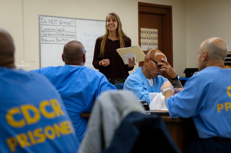 A person stands between four inmates, dressed in blue uniforms with the words "CDCR PRISONER" printed on them in yellow, who are seated at desks. The person holds papers and a manila envelope. The setting is a class setting, inside of a prison.