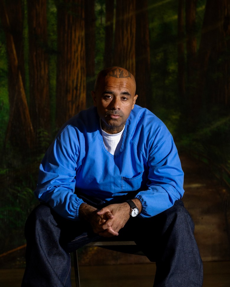 A person with a tattoo on their forehead and wearing a blue shirt sits on a chair, leaning forward with hands clasped. The background shows a forest scene, giving a reflective or contemplative atmosphere.