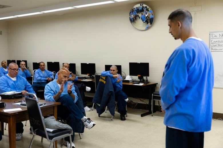 A person dressed in a blue sweatshirt, dressed in black pants, stands in front of a group of people similarly dressed and seated at desks. The setting is a classroom inside of a prison. A row of computers are visible behind the group.