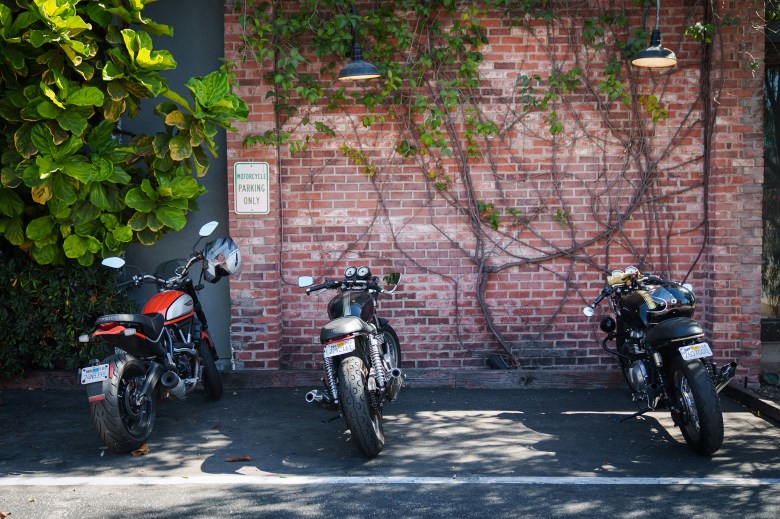 Three motorcycles parked in a parking lot with a motorcycle parking only sign hanging on a brick wall.