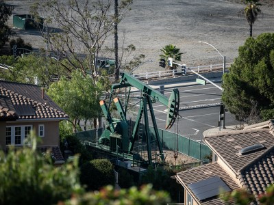 A green active oil pump jack surrounded by two-story homes and a street intersection in the background.