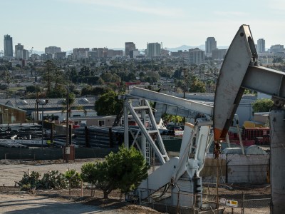 Oil well pumps in the foreground among residential and commercial buildings with a backdrop of the Long Beach skyline in the background.