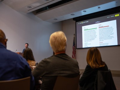 A crowd of people watching a presentation projected on a screen while a man stands and speaks at a podium in a room.