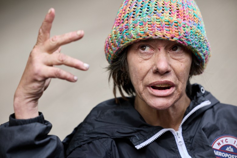 A close-up view of a person wearing a multi-colored beanie hat, a black zip up jacket and golden grilled teeth, as they talk and gesture with their right hand.