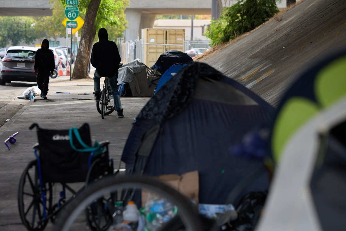 A side view of a man on a bike next to homeless tents lined up along a sidewalk underneath a highway in Sacramento.