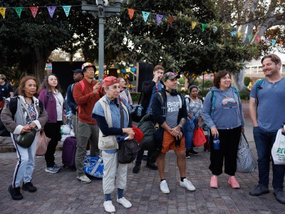 About a dozen people carrying bags, luggage and other personal belongings stand at a public plaza before they board a bus in Los Angeles.