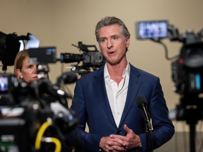 A person in a blue blazer, and an unbuttoned white shirt, stands in front of cameras during a press conference. The view point is from behind the cameras, showing the person at center.