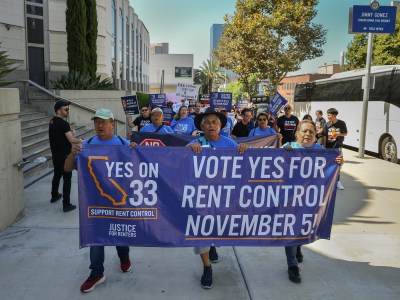 Protestors hold up a banner saying "Vote yes for rent control November 5!" as they lead a march down a sidewalk near a building.