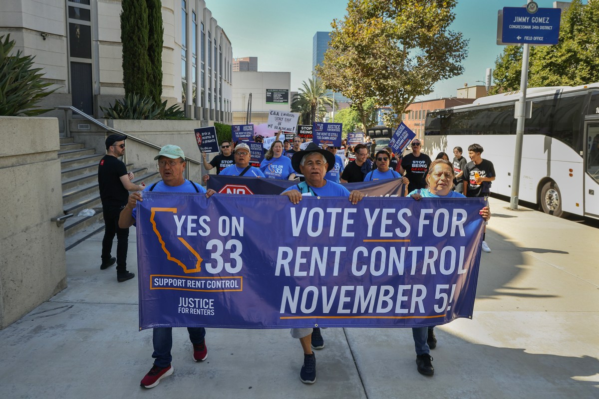 Protestors hold up a banner saying "Vote yes for rent control November 5!" as they lead a march down a sidewalk near a building.