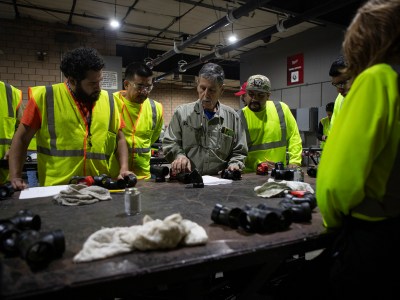 A person is holding a section of pipe while a group of people surrounds him as he teaches a lesson at a table in a workshop.