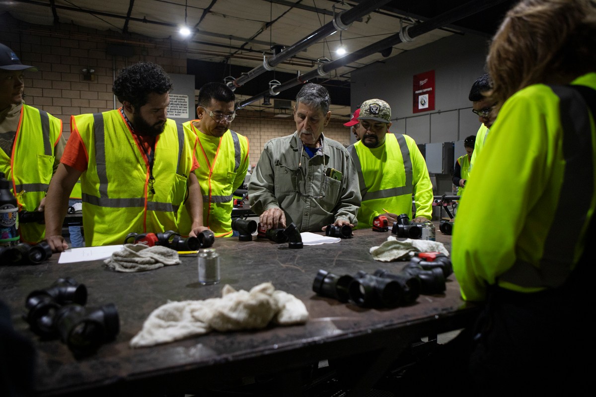 A person is holding a section of pipe while a group of people surrounds him as he teaches a lesson at a table in a workshop.