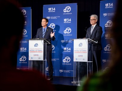 Two men on stage at a podium. The one on the left is talking and gesturing to someone out of frame.