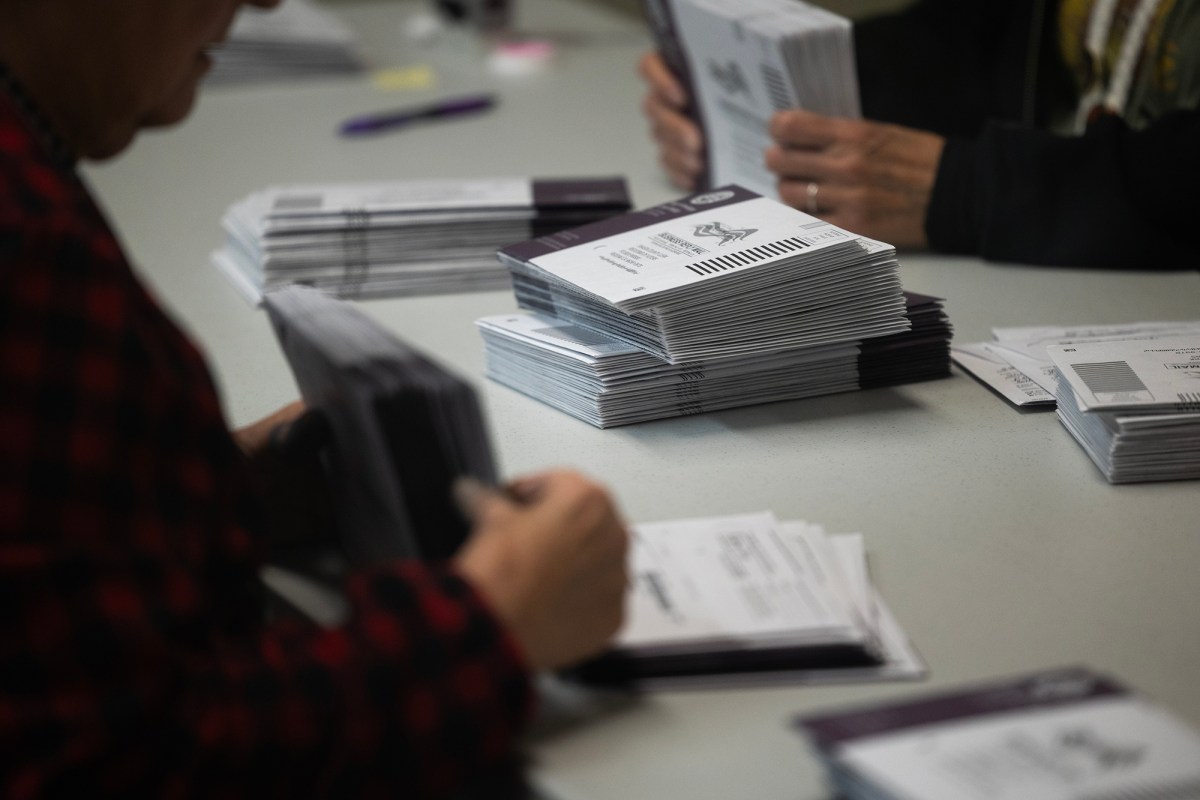 Stacks of purple and white mail-in ballot envelopes sit on a stable as election workers sort them. The arms and hands of the workers are visible, but the faces are not. The focus is on the ballots.