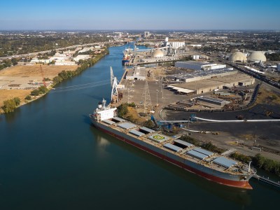 Aerial view of a large cargo ship docked at an industrial port along a wide river. The ship has a helipad marked with an "H" and is equipped with several open cargo holds. Cranes and industrial equipment are visible on the dock, with storage tanks, warehouses, and other infrastructure nearby. Surrounding the port area are open fields, warehouses, and a network of roads, with a cityscape extending into the distance under a clear blue sky.