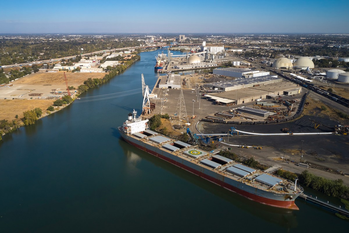 Aerial view of a large cargo ship docked at an industrial port along a wide river. The ship has a helipad marked with an "H" and is equipped with several open cargo holds. Cranes and industrial equipment are visible on the dock, with storage tanks, warehouses, and other infrastructure nearby. Surrounding the port area are open fields, warehouses, and a network of roads, with a cityscape extending into the distance under a clear blue sky.