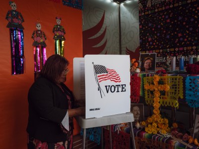 A voter casts a mock-ballot at a voting booth adorned with an American flag and the word "VOTE." The scene is set against a vibrant backdrop, featuring Día de los Muertos decorations and an altar with colorful papel picado, marigolds, and framed photos of loved ones. The cultural ambiance adds depth to the act of civic participation.