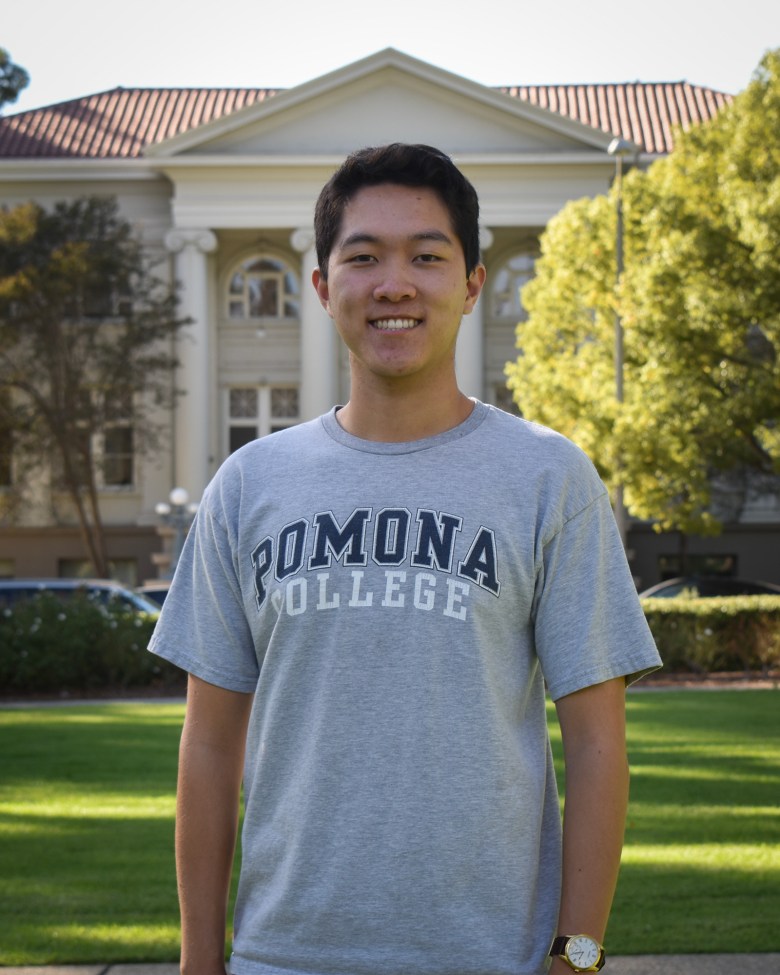 A person wearing a grey shirt with the words "POMONA COLLEGE," stands with their arms to their sides in front of building with Greco-Roman columns at front. The scene evokes a college campus.